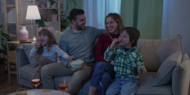 Dad showing something funny on the TV. Family sitting on the sofa eating popcorn and laughing while having a movie night
