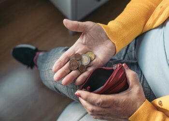 Close-up aerial image of a seated older woman's hand holding coins in her palm while holding a purse in her other hand.